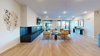 Located at 705 1st Ave SW in Rochester, Minnesota, this clubroom and sports simulator facility offers a well-appointed, single-floor amenity space designed for both relaxation and active recreation. The main lounge area is generously sized, featuring warm light wood flooring, large black-framed windows that invite abundant natural light, and a striking dark-tiled fireplace flanked by a wall-mounted television. Comfortable seating arrangements with contemporary furnishings create an inviting social atmosphere throughout the open layout. Adjacent to the lounge is a sleek kitchenette outfitted with white cabinetry, white quartz countertops, a stainless steel sink with a gold-tone faucet, and a bold navy blue vertical tile backsplash that adds a vibrant design focal point. Open metal shelving and a built-in microwave complete the functional space. The sports simulator room is a standout feature, housing a Full Swing simulator with a large projection screen, dark carpet turf flooring, and ample space for equipment storage. A dedicated viewing and lounge area adjacent to the simulator includes patterned carpet, a long communal table, and curated wall art with a golf theme, providing a comfortable space to watch and socialize.