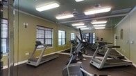 This fitness center is a dedicated single-floor workout facility designed to support a well-rounded exercise routine. The room is outfitted with a suite of Matrix-brand equipment, including multiple treadmills, elliptical cross-trainers, and a multi-station cable and weight machine, providing users with comprehensive cardio and strength training options. The space is finished with dark, durable flooring suited for high-traffic use and is painted in a muted olive green, lending the room a focused, energetic atmosphere. Mirrored walls along one side visually expand the space and allow users to monitor their form during workouts. Bright overhead fluorescent lighting ensures the entire floor is evenly and thoroughly illuminated at all times. Several windows fitted with horizontal blinds line the walls, offering the option for natural light and ventilation. Ceiling fans are installed to assist with air circulation, contributing to a more comfortable workout environment. The layout is thoughtfully arranged to allow easy movement between machines, making the facility accessible and functional for multiple users simultaneously.