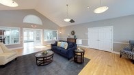 This single-story space is thoughtfully designed to balance comfort and functionality across its open layout. The main gathering area commands attention with soaring vaulted ceilings and a striking stone fireplace flanked by large windows that flood the room with natural light. An arched transom window above the fireplace adds architectural character, while pendant lighting and recessed fixtures ensure warm, even illumination throughout. Hardwood flooring in a light honey tone runs seamlessly through the entire space, enhancing the sense of continuity and openness.

The kitchen is well-appointed with rich cherry cabinetry featuring raised-panel doors, dark granite countertops, and a subway tile backsplash. A central island with a black granite surface provides additional workspace and bar seating, complementing the black appliances including a refrigerator, range, and built-in microwave. A dedicated sink area with ample lower cabinet storage rounds out the practical design.

Adjacent to the kitchen, an open sitting area offers flexible seating arrangements with generous wall space and large windows overlooking the surrounding greenery. The neutral gray wall tones, white trim, and chair-rail detailing throughout lend the space a clean, polished aesthetic suitable for a variety of uses.