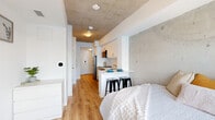 Located at 575 Princess Street in Kingston, Ontario, this studio unit offers a thoughtfully designed open-concept layout that maximizes every square foot. The space is defined by its striking exposed concrete ceiling, which adds an industrial-modern character throughout, complemented by warm light wood flooring and crisp white walls and cabinetry.

The kitchen runs along one side of the unit and is equipped with stainless steel appliances, including a refrigerator, stove, and dishwasher, alongside white subway tile backsplash and ample upper cabinetry. A peninsula counter with bar stools provides a casual dining and workspace area, seamlessly connecting the kitchen to the sleeping area. The living and sleeping space is brightened by a large window with city views, and includes a dedicated study area with a wall-mounted television.

A full bathroom features a clean, minimalist design with a white vanity, large mirror, and tiled flooring. A practical in-unit closet conceals a stacked washer and dryer, adding everyday convenience. The unit also benefits from a smart lock entry system and a modern thermostat. Overall, the space delivers a functional, contemporary living environment well-suited for students or young professionals in downtown Kingston.
