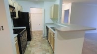 This single-story space presents a practical and functional layout anchored by a well-equipped kitchen. The kitchen features light wood cabinetry both above and below the countertops, providing ample storage throughout. Black appliances, including a refrigerator and range, offer a subtle contrast against the neutral-toned countertops and white walls. A double-basin sink is neatly integrated into the counter, positioned alongside a dishwasher for added convenience. A peninsula counter extends outward, creating a natural division between the kitchen and the adjoining area, and could serve as a casual dining or prep space. The flooring throughout features stone-look tile in warm, earthy tones, adding texture and durability to the space. A pass-through window on the far wall allows for visual connectivity to an adjacent room, enhancing the open feel of the layout. Just off the kitchen, a spacious walk-in closet is visible, offering generous storage capacity. The overall ambiance is clean and straightforward, with a neutral color palette of whites and beiges that creates a bright, airy atmosphere throughout the space.