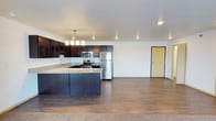 This single-floor unit showcases a thoughtfully designed open-concept layout that seamlessly connects the kitchen and living areas, creating a spacious and functional environment. The kitchen is a standout feature, highlighted by rich dark espresso cabinetry that extends to the upper walls, offering generous storage. Light granite countertops provide a striking contrast, complemented by a kitchen island with a built-in sink — ideal for meal preparation and casual dining. Stainless steel appliances, including a refrigerator, range, and over-the-range microwave, add a sleek, contemporary finish. A modern pendant light fixture hangs above the island, providing both task lighting and a decorative focal point. Recessed lighting throughout the ceiling ensures the space remains bright and well-illuminated. The flooring transitions from warm hardwood-style planks in the kitchen and main living area to plush carpet toward the entry, adding comfort and texture. The open living area provides ample room for a variety of furniture arrangements. The unit's entry door is clearly defined, offering a clean transition from the exterior. The overall ambiance is modern, clean, and move-in ready, appealing to those who value both style and practicality.