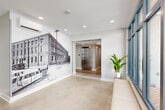 Located at 1717 Religious St in the heart of New Orleans, this building hallway serves as a welcoming and well-appointed entry point that seamlessly merges historical character with contemporary design. The space is defined by polished concrete floors that extend throughout, lending an industrial yet refined aesthetic that anchors the overall ambiance.

A standout feature is the large-scale black-and-white photographic mural depicting the building's historic identity as the Seelig Furniture Manufacturing Co., serving as a visual centerpiece that pays tribute to the property's rich past. This artwork wraps dramatically across a prominent interior wall, immediately capturing the attention of anyone entering the space.

Floor-to-ceiling windows fitted with dark-framed mullions line one side of the hallway, inviting generous natural light and offering views to the exterior streetscape. A built-in upholstered bench in a textured blue fabric provides a functional seating area beneath the windows, complemented by a potted tropical plant that adds a fresh, organic touch.

Recessed ceiling lighting ensures the space is well-lit throughout, while a wall-mounted mini-split HVAC unit provides climate control. A stainless steel elevator is accessible from within the hallway, connecting to the broader building. The entry vestibule features a floor-level entry mat and a glass-paneled door that opens directly to the street, ensuring a clean and functional transition between interior and exterior.