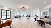 Located at 3100 Purcell St in Richmond, Virginia, this single-floor community space offers a thoughtfully designed open-concept layout combining a kitchen and living room. Light hardwood flooring runs throughout, lending warmth and visual continuity across the entire space.

The living area is divided into distinct seating zones, including a lounge area near the entrance appointed with cognac leather armchairs arranged atop a geometric area rug beneath a striking circular pendant light fixture. A secondary seating nook features a gray sofa paired with an accent chair set against a bold teal accent wall, adding a vibrant pop of color to the otherwise neutral palette. Multiple dining tables with modern chairs provide flexible gathering options throughout the open floor plan.

Large windows along the perimeter bathe the interior in natural light and offer views of the surrounding greenery. A welcoming set of glass double doors at the entrance connects the space to the outdoors.

The kitchen area features rich dark wood cabinetry with brushed metal hardware, a white quartz countertop, a stainless steel sink, and a built-in microwave. Open floating shelves mounted above a textured white tile backsplash add both style and function, while a full-size refrigerator completes the setup. Recessed lighting throughout ensures even illumination across all areas of this versatile, well-appointed space.
