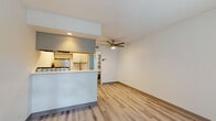 This single-floor space offers a clean, modern aesthetic defined by light wood-style flooring that runs consistently throughout every room, creating a seamless and cohesive feel. The open-concept living and kitchen area serves as the heart of the unit, where a peninsula-style kitchen counter separates the two spaces while maintaining an airy, connected atmosphere. The kitchen is thoughtfully designed with white cabinetry, quartz-style countertops, a gas range, a refrigerator, and an overhead ventilation hood — all set within a galley-style configuration that maximizes efficiency. A ceiling fan with a built-in light fixture anchors the living area, providing both comfort and illumination. The entryway welcomes natural light through a large window fitted with blinds alongside a modern front door with glass panel inserts, lending the space a bright and inviting first impression. The bathroom features a white vanity with a quartz countertop, a framed mirror with warm-toned accents, and tile wall detailing, offering a clean and updated finish. White walls throughout the space enhance the sense of brightness and openness, while consistent trim and hardware details tie the overall design together in a fresh, contemporary style.