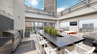 Located in the heart of Downtown Los Angeles, this rooftop amenity deck at 225 S Grand Ave offers a compelling blend of open-air and semi-enclosed spaces designed for both relaxation and social gatherings. The deck is framed by floor-to-ceiling glass panels that provide sweeping, unobstructed views of the downtown LA skyline and surrounding cityscape, flooding the space with abundant natural light throughout the day.

The layout is thoughtfully divided into several distinct functional zones. A fully equipped outdoor kitchen anchors one section, featuring dual stainless steel gas grills, under-counter refrigeration units, a sink, and dark stone countertops set against a sleek tile backsplash. Adjacent to the kitchen is a large bar-height communal table with built-in planters and white bar stools, ideal for dining and entertaining.

Multiple lounge areas are furnished with plush, neutral-toned sectional sofas and wicker coffee tables, offering comfortable spots to unwind while taking in the city views. A standalone fireplace with surrounding seating provides a cozy focal point for the open-air terrace section. A dedicated dining area with a white table and vibrant orange chairs adds a lively pop of color to the space. Wall-mounted flat-screen televisions and overhead string lights enhance the ambiance, making this rooftop deck a versatile, amenity-rich retreat above the city.