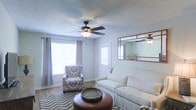Located at 1000 Vineyard Dr in Brandon, Mississippi, this single-story home offers a comfortable and inviting living environment designed for easy, functional living. The living room serves as the heart of the home, showcasing a light and airy atmosphere enhanced by large windows dressed with gray curtains that allow generous amounts of natural light to fill the space. A classic ceiling fan with a built-in light fixture is centrally mounted, providing both comfort and illumination. The room features neutral-toned walls that complement the warm hardwood-style flooring, creating a cohesive and welcoming aesthetic. A decorative wall mirror with a warm-toned frame adds depth and visual interest to the space. The layout comfortably accommodates a full sofa, accent chair, and a round ottoman, making it well-suited for relaxing or entertaining. The overall ambiance of the home is calm and understated, with a clean, move-in-ready feel. Its single-floor layout offers convenient accessibility throughout, and its location in Brandon, MS places it within a well-established residential community.