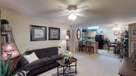 This single-floor residence showcases an open-concept floor plan that seamlessly connects the living area to the kitchen and dining space. The living room features plush, wall-to-wall carpeting in a neutral beige tone, complemented by warm-colored walls that create a welcoming atmosphere. A ceiling fan with integrated lighting serves as a functional focal point overhead, while two framed botanical artworks add a decorative touch to the main wall. A dark gray sofa paired with a classic wooden coffee table anchors the seating area.

The space flows naturally into a compact yet well-equipped kitchen, which features light wood cabinetry, a black refrigerator, and a breakfast bar with two bar-height chairs — ideal for casual dining. Under-cabinet lighting illuminates the countertops, enhancing the kitchen's functionality. A pass-through design between the living and kitchen areas promotes an open, airy feel throughout the home.

A hallway visible from the living room suggests access to additional private rooms, and a decorative floor lamp near the transition area adds a warm, inviting glow. Overall, the space presents a cohesive, comfortable layout suited for everyday living.