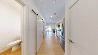 Located at 399 S Crouse Ave in Syracuse, NY, this efficiently designed unit offers a thoughtful layout across a single floor. The open corridor connects all areas seamlessly, with warm-toned luxury vinyl plank flooring running throughout the space, contributing to a clean, cohesive aesthetic.

The kitchen is well-appointed with stainless steel appliances, including a French door refrigerator, electric range, over-the-range microwave, and dishwasher. Light wood upper cabinetry is paired with white lower cabinets and a light-toned countertop, creating a modern and inviting cooking area. A stainless steel sink with a contemporary faucet adds a functional touch.

Adjacent to the kitchen, a pantry area provides additional storage, accessible via a sleek sliding barn door — a stylish detail that adds character to the space. The hallway also features an electrical panel recessed into the wall for convenience.

The bathroom is clean and modern, featuring a spacious walk-in shower with white surround walls, a rainfall showerhead, and chrome fixtures. A vanity with mirror and toilet complete the well-organized bathroom.

The bedroom area is bright and airy, highlighted by a large window that fills the room with natural light and offers views of the surrounding neighborhood. The light walls and flooring enhance the open feel of this compact yet functional space.