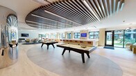 Located at 377 Crane St in Orange, NJ, this single-floor amenity space is designed with a contemporary aesthetic that seamlessly blends recreation, socializing, and productivity. The expansive game lounge serves as a central hub, featuring multiple pool tables set beneath a striking slatted wood ceiling with integrated linear lighting and large windows that invite natural light throughout. Transitioning from the game area, the open social lounge offers generous seating arrangements with modern sofas, vibrant accent chairs in bold orange and yellow, and a sleek fireplace topped by a wall-mounted TV — all set atop wide-plank hardwood flooring. A stylish bar counter with white quartz surfaces and warm gold cabinetry runs along one side of the space, complemented by leather-upholstered barstools and additional mounted displays. The well-appointed kitchen features extensive cabinetry, a large island with a sink, and abundant counter space. Private meeting and work rooms are also available, each equipped with built-in desks, wall-mounted screens, and acoustic wall panels for a focused environment. Throughout the space, curated artwork, recessed and pendant lighting, and glass partitions contribute to an overall ambiance that is polished, inviting, and thoughtfully designed for community living.