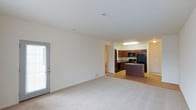 This 2-bedroom, 2-bathroom single-floor residence offers a thoughtfully designed open-concept layout that maximizes both space and functionality. The living area features neutral-toned carpeting and light-colored walls that create a bright, welcoming atmosphere. A sliding glass door with blinds allows natural light to fill the room and provides easy access to the outdoors.

The kitchen is seamlessly integrated into the living space, featuring rich dark wood cabinetry, a central island with a breakfast bar, and black appliances including a refrigerator, microwave, and range. The kitchen transitions to hard surface flooring, offering a clean contrast to the carpeted living area. Recessed and overhead lighting ensures the kitchen is well-lit for meal preparation.

The open sightlines between the living and kitchen areas make this floor plan ideal for entertaining or everyday living. The neutral color palette throughout provides a versatile backdrop for a variety of interior design styles. With two full bathrooms and two bedrooms, this residence delivers a comfortable and practical living experience suited to a range of lifestyles.