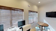 Located at Dozier, this single-floor space offers a clean, well-lit environment designed for both productivity and comfort. The interior is characterized by crisp white walls and recessed lighting that work together to create a bright, inviting ambiance throughout. Large windows dressed with warm-toned bamboo blinds allow natural light to filter softly into the room while maintaining a sense of privacy. The flooring is light-toned and easy to maintain, complementing the overall neutral color palette of the space. A dedicated workstation area is equipped with a desktop computer setup, making it well-suited for administrative or professional use. Adjacent to the workstation, a casual seating area features a rustic wooden high-top table paired with light blue metal stools, adding a touch of character and color to the space. The ceiling features a drop-tile design with integrated recessed lighting and a security camera, reflecting a practical and functional approach to the layout. The combination of natural light, thoughtful furnishings, and a tidy, organized atmosphere makes this space versatile and well-appointed for a variety of uses. The overall design strikes a balance between functionality and approachability, making it a comfortable environment for visitors and occupants alike.