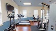 Located at 2220 Gloria Cir in Pensacola, Florida, this single-floor fitness center is designed to provide a well-rounded exercise environment. The space features warm hardwood flooring throughout, complemented by a two-tone interior with soft blue wainscoting and cream-colored upper walls accented by crown molding, giving the room a clean and polished aesthetic. Multiple windows fitted with white blinds allow generous natural light to fill the space, creating a bright and energizing atmosphere. A large wall mirror spans one section of the room, visually expanding the space and allowing users to monitor their form during workouts. The facility is equipped with a comprehensive selection of exercise equipment, including Spirit-brand treadmills, a recumbent stationary bike, a rowing machine, an elliptical trainer, and a multi-station cable weight machine. Protective rubber floor mats are positioned beneath the equipment for safety and durability. Overhead fluorescent lighting ensures consistent illumination throughout all areas. A mounted flat-screen television is available for entertainment during cardio sessions. French doors with glass panels provide access to the exterior, adding additional natural light and a connection to the outdoors. The overall layout is thoughtfully organized to maximize both functionality and comfort for users.