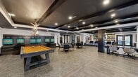Located at 401 E Oglethorpe Ave in Savannah, Georgia, this expansive single-floor clubroom delivers a sophisticated social environment designed for both relaxation and entertainment. The open-concept layout is anchored by a dramatic coffered ceiling finished in deep navy and black, accented by exposed dark wood beams and recessed lighting that creates a warm, upscale ambiance throughout.

The space is thoughtfully divided into distinct activity zones. A standout pool table with an amber felt top serves as the centerpiece of the entertainment area, flanked by leather lounge seating and vibrant teal-accented wall art. Nearby, round dining tables with modern chairs offer a casual gathering spot. A striking full-length bar counter clad in veined marble stone is lined with sleek upholstered barstools and pendant lighting, complemented by a well-appointed back bar with display shelving.

A dedicated workspace with built-in desk surfaces and rolling chairs provides a functional area for productivity. Large French-style glass doors along multiple walls allow natural light to filter in and offer views of the outdoor surroundings. The durable tile flooring, dark structural columns, and curated artwork tie the entire space together, resulting in a refined yet inviting clubroom atmosphere.