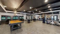 Located at 401 E Oglethorpe Ave in Savannah, Georgia, this expansive single-floor clubroom delivers a sophisticated social environment designed for both relaxation and entertainment. The open-concept layout is anchored by a dramatic coffered ceiling finished in deep navy and black, accented by exposed dark wood beams and recessed lighting that creates a warm, upscale ambiance throughout.

The space is thoughtfully divided into distinct activity zones. A standout pool table with an amber felt top serves as the centerpiece of the entertainment area, flanked by leather lounge seating and vibrant teal-accented wall art. Nearby, round dining tables with modern chairs offer a casual gathering spot. A striking full-length bar counter clad in veined marble stone is lined with sleek upholstered barstools and pendant lighting, complemented by a well-appointed back bar with display shelving.

A dedicated workspace with built-in desk surfaces and rolling chairs provides a functional area for productivity. Large French-style glass doors along multiple walls allow natural light to filter in and offer views of the outdoor surroundings. The durable tile flooring, dark structural columns, and curated artwork tie the entire space together, resulting in a refined yet inviting clubroom atmosphere.