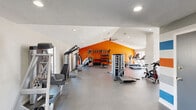 The Fitness Center at 1400 Sierra Springs Dr in Bedford, Texas is a well-equipped, single-floor exercise facility designed to support a comprehensive workout experience. The generously sized exercise room features vaulted ceilings that amplify the sense of openness, complemented by recessed lighting and a ceiling fan that help maintain a comfortable environment during workouts. A textured ceiling adds a subtle architectural detail throughout the space.

The facility is organized to accommodate a full range of training modalities. The cardio zone is equipped with multiple treadmills and incline trainers, positioned near large windows and glass double doors that open directly to an outdoor pool area, flooding the space with natural light and providing pleasant exterior views. The strength training area is anchored by a bold orange accent wall, which serves as a vibrant focal point and houses a well-stocked wall-mounted rack of kettlebells and free weights. Multiple cable machines, a multi-use functional trainer, weight benches, and selectorized strength machines are thoughtfully arranged to maximize usable floor space. Cycling bikes round out the cardio offerings. Durable dark flooring runs throughout, providing a practical and sleek surface suited for high-traffic use. Informational signage is displayed near the entrance, and the overall color palette of white, orange, and blue gives the center a modern, energetic aesthetic.