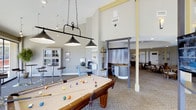 Located at 890 Quail Lake Circle in Colorado Springs, Colorado, this community clubhouse offers a thoughtfully designed single-floor gathering space built for recreation and relaxation. The centerpiece of the main room is a full-size billiards table, elegantly illuminated by a classic hanging pendant light fixture suspended from soaring vaulted ceilings that lend an open, airy feel to the space. Neutral-toned carpeting and soft, recessed lighting throughout create a comfortable and welcoming ambiance. A dedicated lounge area features plush seating arranged around a flat-screen television, while a separate nook provides a quiet workspace complete with a desk and chair. The clubhouse also includes a wet bar or service counter area with a curved, metallic-accented design, adding a modern touch to the interior. A dining area with ample seating is visible through a connecting passage, making the space well-suited for community gatherings. Large sliding glass doors along one wall flood the interior with natural light and offer direct access to an outdoor patio or deck, where additional seating and lush greenery can be found. The overall design balances functionality with style, providing residents with a versatile space for both leisure and socializing.