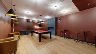 This single-floor amenity lounge is a well-designed recreational space that combines entertainment and relaxation in a cohesive layout. The room is unified by deep terracotta-red walls and light-toned hardwood flooring, creating a warm yet energetic ambiance throughout. A full-size pool table with a red felt surface serves as the centerpiece, positioned beneath a set of industrial-style pendant lights that provide focused illumination. A built-in electric fireplace framed by a teal textured accent column adds a striking architectural focal point along one wall.

Bar-height tables and stools line the perimeter, offering casual seating for socializing. A dedicated lounge corner features comfortable sofas, a wall-mounted flat-screen television, wall-mounted cue racks, and decorative shelving. Additional entertainment options include arcade game cabinets near the entry. The entrance area features glass-paneled doors, black trim detailing, and a leather accent chair, contributing to the polished overall design.

Recessed and pendant lighting fixtures are distributed throughout the ceiling, ensuring consistent, well-layered illumination across the entire space. The combination of recreational amenities, thoughtful furnishings, and bold interior design makes this lounge a versatile and inviting communal gathering space.