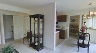 This single-floor residence offers a thoughtfully arranged open layout that connects the main living area, kitchen, and bedroom in a cohesive, functional flow. The living area is finished with plush carpeting in a neutral tone, creating a warm and inviting atmosphere. A decorative display cabinet with glass panels and dark wood framing serves as a charming focal point, accompanied by a classic accent chair that adds a touch of traditional elegance to the space.

A structural column partially divides the living area from the kitchen, providing a sense of definition without fully closing off the space. The kitchen features warm wood-toned cabinetry, a light-colored countertop with a breakfast bar overhang, and a clean, efficient layout. The cabinetry provides ample storage, and the neutral wall tones keep the kitchen feeling bright and open.

Beyond the kitchen, a glimpse into the bedroom reveals continued carpeting and a striped bedding set, suggesting a comfortable and consistently styled private retreat. The overall color palette throughout the space is understated and cohesive, combining beige walls, warm wood tones, and soft flooring to create a calm, livable environment. The single-floor plan ensures convenient, accessible navigation throughout the home.