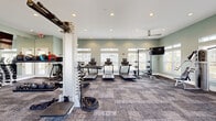 Located at 1110 Mason Pond Pl in Ladson, SC, this fitness center offers a generously sized, single-floor layout designed to accommodate a comprehensive range of workout activities. The space is divided into two functional areas, each thoughtfully appointed to serve different training needs.

The main fitness floor is bathed in natural light streaming through numerous large windows fitted with white blinds, complemented by recessed ceiling lighting and ceiling fans that maintain a comfortable environment. Soft sage-green walls and neutral carpet tile flooring create a clean, inviting atmosphere. This area houses a full array of cardio equipment, including treadmills, elliptical machines, and a rowing machine, alongside a multi-station cable machine, a power rack with bench, a weight machine, and various free weights and functional training accessories.

A dedicated secondary space serves as a spin studio, featuring a row of stationary cycling bikes positioned before a floor-to-ceiling mirror wall. A wall-mounted flat-screen TV enhances the workout experience, and tall windows allow natural light to fill the room. A water cooler station is conveniently available nearby. Throughout the facility, the design emphasizes openness, functionality, and a bright, motivating ambiance well-suited for residents seeking a complete on-site fitness experience.