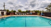 Located at 5 Mooney Rd NE in Fort Walton Beach, Florida, this outdoor amenity space centers around a generously sized rectangular swimming pool with crystal-clear turquoise water. The pool is equipped with stainless steel entry rails and is clearly depth-marked for safety. Surrounding the pool is a spacious concrete sundeck furnished with rows of teal lounge chairs, ideal for relaxing in the Florida sunshine. A covered gazebo sits at one corner of the space, outfitted with ceiling fans and additional seating, providing a shaded retreat for residents and guests. A dedicated green lawn area completes the outdoor layout, featuring a grill station and casual seating, perfect for outdoor gatherings. Dining-height tables with chairs are also positioned poolside for convenience. The entire pool area is enclosed by a combination of iron fencing and a wooden privacy fence, creating a secure and semi-private environment. Mature trees along the perimeter offer natural shade and a lush backdrop. The adjacent multi-story building with balconies is visible from the pool area, reinforcing the community-oriented nature of this well-appointed outdoor space. Overall, the area is designed to deliver a resort-like experience with a variety of amenities suited for both relaxation and socializing.