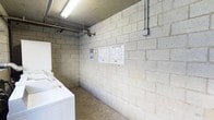 This single-floor laundry facility is designed with functionality and durability at its core. The room features exposed concrete block walls that give the space an industrial character, complemented by a polished concrete floor that is easy to maintain. Overhead, fluorescent strip lighting provides bright, even illumination throughout the area, ensuring excellent visibility for users at any time of day. Exposed conduit and utility pipes run along the upper walls and ceiling, reinforcing the utilitarian nature of the space. The facility is equipped with white top-loading washing machines positioned along one wall, making efficient use of the available floor space. Operating instruction placards are clearly posted on the wall, offering users straightforward guidance on machine usage. A coin-operated payment unit is also visible, indicating this is a shared, pay-per-use laundry space. The overall layout is straightforward and practical, prioritizing ease of access and efficient workflow for laundry tasks. The no-frills design ensures that the space is easy to clean and maintain, making it a reliable utility area suited for shared residential or commercial use.