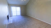 This single-floor space is defined by its vaulted ceilings, which lend a sense of openness and airiness to the main living area. The soaring roofline draws the eye upward, creating a feeling of generous vertical space that enhances the overall roominess of the interior. A ceiling fan is mounted at the peak, providing both function and a subtle design element that complements the angular ceiling lines.

The flooring throughout consists of plush wall-to-wall carpeting in a neutral beige-gray tone, offering a soft and comfortable underfoot experience while contributing to the room's understated, cohesive aesthetic. Neutral-toned walls in a warm greige shade provide a versatile backdrop suitable for a variety of décor styles.

A prominent sliding glass door, dressed with vertical blinds, serves as a key architectural feature of the space. It allows an abundance of natural light to filter into the room, brightening the interior throughout the day while also providing direct access to an outdoor area. White baseboards frame the perimeter of the room cleanly, adding a crisp finishing detail. Overall, the space presents a calm, open, and welcoming atmosphere.