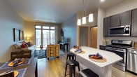This single-floor residence offers a thoughtfully designed open-concept layout that seamlessly connects the kitchen, dining, and living areas into one cohesive living space. The kitchen is appointed with dark cabinetry, granite countertops, and a breakfast bar overhang that accommodates bar-height seating — ideal for casual dining or entertaining. A trio of pendant lights hangs above the bar, providing both task lighting and a stylish focal point. The flooring throughout the main living area is warm hardwood, adding a sense of continuity and natural warmth. The living area is generously proportioned, bathed in natural light streaming through large floor-to-ceiling windows that frame views of an outdoor balcony. The balcony access enhances the sense of openness and provides a pleasant indoor-outdoor connection. Neutral gray walls provide a versatile backdrop, complemented by warm wood-toned door frames and trim. A hallway visible from the main area leads to additional rooms, suggesting a practical and well-organized floor plan. The overall ambiance is contemporary and welcoming, balancing modern finishes with a comfortable, livable aesthetic suited for a variety of lifestyles.