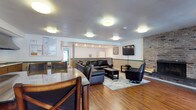 This single-floor space is thoughtfully designed for communal gathering, combining comfort and functionality throughout its well-appointed areas. Rich hardwood flooring runs continuously across the entire space, lending warmth and visual continuity to every room.

The central lounge area serves as the heart of the space, anchored by a dramatic floor-to-ceiling exposed brick fireplace that adds timeless character and charm. Comfortable black leather seating — including a sectional sofa, a tufted ottoman, and a swivel accent chair — is arranged around a soft area rug, creating an inviting setting for relaxation. A wall-mounted flat-screen television completes the entertainment setup.

Adjacent to the lounge, a raised dining area accommodates a large high-top table with six cushioned chairs, ideal for group meals or meetings. Windows and a classic green Dutch door provide natural light and views of the surrounding brick exterior, enhancing the sense of connection to the outdoors.

A compact kitchenette supports the space with essential amenities, including a countertop cooktop, sink, cabinetry, and a ventilation hood. A small table and chair offer an additional informal seating option nearby.

Throughout the space, flush-mount ceiling lights provide consistent, even illumination. A green accent stripe along the walls ties the rooms together with a subtle, cohesive design touch.