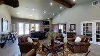 This single-floor space exudes rustic charm and warmth, thoughtfully designed with a cohesive blend of natural materials and inviting details. The main living area commands attention with soaring vaulted ceilings accented by an exposed dark wood beam, creating a sense of openness and architectural character. Recessed lighting throughout ensures a well-lit interior, while large French doors along one wall flood the space with abundant natural light and provide views to an outdoor area beyond.

A striking floor-to-ceiling stone fireplace serves as the room's focal centerpiece, topped with a mounted television and flanked by decorative accents on the mantle. Rich, brown leather seating is arranged around an ornate wooden coffee table atop a detailed area rug, fostering a comfortable and inviting gathering environment. A sliding barn door with classic hardware adds a distinctive rustic touch and leads to an adjacent room. Additional wooden furniture and cabinetry throughout reinforce the warm, lodge-inspired aesthetic. The overall ambiance of the space is one of relaxed sophistication — combining natural textures, thoughtful design elements, and a neutral color palette to create a welcoming and functional environment.