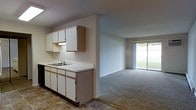 This single-story space features an open-concept layout that connects the kitchen and living area seamlessly. The kitchen is equipped with white cabinetry, both upper and lower, accented by light wood trim, and includes a double-basin sink set into a light-colored countertop. Tile flooring grounds the kitchen area, while the adjacent living space transitions to wall-to-wall carpet, creating a distinct yet cohesive separation between the two zones. The living area is bright and open, benefiting from a large sliding glass door that fills the room with natural light and provides a view of a grassy outdoor area. The neutral wall tones throughout contribute to a clean, understated aesthetic. The overall layout is practical and functional, offering an easy flow between cooking and living spaces, making it well-suited for everyday use.