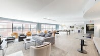 This expansive single-floor amenity space is thoughtfully designed to offer a diverse range of social and recreational experiences within one open, flowing layout. Light wood-look flooring runs throughout, unifying the various zones while contributing to the bright, airy atmosphere created by an abundance of large, floor-to-ceiling windows along the perimeter walls.

The main lounge area is anchored by a striking geometric feature wall clad in textured gray panels, providing a dramatic backdrop for a mounted flat-screen display and a floating media shelf styled with sculptural décor. Comfortable seating arrangements invite relaxation, while the open plan seamlessly connects to the games area, which centers on a full-size billiards table illuminated by a statement gold pendant fixture. Adjacent seating clusters with amber-toned armchairs make the most of the natural light pouring in through the expansive windows.

The gourmet kitchen island serves as a social hub, featuring a generous white quartz countertop, stylish leather barstools with gold accents, pendant lighting, a built-in beverage cooler, and curated wall art. A separate lounge corridor features an oversized decorative mirror, upholstered banquette seating, and bistro-style tables, offering a more intimate setting. Glass double doors provide a welcoming entry point, completing this well-appointed communal space.