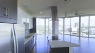 This single-floor space showcases a sleek, contemporary kitchen designed with both style and function in mind. The most striking architectural feature is the wraparound floor-to-ceiling glass windows that flood the space with abundant natural light while offering expansive panoramic views of the surrounding cityscape and tree canopy below, creating an immersive connection with the urban environment outside.

The kitchen is anchored by a large central island topped with light stone countertops, providing ample workspace and casual seating. A bold, oversized stainless steel island range hood serves as a dramatic focal point, drawing the eye upward toward the high ceilings and reinforcing the industrial-modern aesthetic. Cabinetry along the perimeter features a two-tone design, combining crisp white upper cabinets with darker lower units, complemented by matching stone countertops and a built-in sink. High-end stainless steel appliances, including a refrigerator and dishwasher, are seamlessly integrated into the design. Rich dark hardwood flooring grounds the space, adding warmth that contrasts beautifully with the cool tones of the cabinetry and metallic accents throughout.