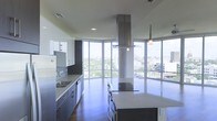 This single-floor space showcases a sleek, contemporary kitchen designed with both style and function in mind. The most striking architectural feature is the wraparound floor-to-ceiling glass windows that flood the space with abundant natural light while offering expansive panoramic views of the surrounding cityscape and tree canopy below, creating an immersive connection with the urban environment outside.

The kitchen is anchored by a large central island topped with light stone countertops, providing ample workspace and casual seating. A bold, oversized stainless steel island range hood serves as a dramatic focal point, drawing the eye upward toward the high ceilings and reinforcing the industrial-modern aesthetic. Cabinetry along the perimeter features a two-tone design, combining crisp white upper cabinets with darker lower units, complemented by matching stone countertops and a built-in sink. High-end stainless steel appliances, including a refrigerator and dishwasher, are seamlessly integrated into the design. Rich dark hardwood flooring grounds the space, adding warmth that contrasts beautifully with the cool tones of the cabinetry and metallic accents throughout.