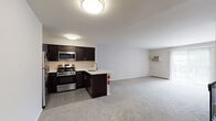 Located at 275 E High St in Glassboro, New Jersey, this single-floor residential unit offers a well-designed layout with a seamless flow between the kitchen and living areas. The kitchen is a standout feature, equipped with rich dark espresso cabinetry, stainless steel appliances including a gas range, microwave, refrigerator, and dishwasher, all complemented by light countertops and tile flooring. A peninsula counter separates the kitchen from the open living area, which is finished with neutral wall-to-wall carpeting and includes a wall-mounted air conditioning unit and baseboard heating. Natural light filters in through sliding glass doors leading to a balcony-style access point, enhancing the airy feel of the living space. The bedroom is a comfortable, carpeted room with a window providing natural light and its own wall unit for climate control. The bathroom features a tiled tub-shower combination with a classic black-and-white tile accent border, a window for ventilation, and clean, functional fixtures. A staircase with a black metal railing is also visible within the unit, indicating shared building access. Overall, the space offers a clean, modern aesthetic with practical finishes throughout.
