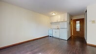 This single-floor unit offers a straightforward, functional layout designed for efficient everyday living. The main living area is open and uncluttered, featuring rich dark hardwood-style flooring that extends throughout the space, creating a cohesive and grounded aesthetic. Neutral cream-toned walls provide a clean, bright backdrop, while baseboard trim adds a classic finishing touch.

The kitchenette is neatly integrated into the living area and includes white shaker-style upper cabinetry offering ample storage, a stainless steel sink with a single-basin basin, a white gas range, and a full-size top-freezer refrigerator. A closet with a hanging rod is conveniently positioned adjacent to the kitchen area, providing additional storage options.

The bedroom is a calm, well-proportioned room bathed in natural light from a window overlooking lush greenery. Dark hardwood-style flooring continues here, and a baseboard heating unit runs along the exterior wall, ensuring comfortable warmth throughout cooler seasons.

The bathroom features a clean, white-tiled tub and shower combination with chrome fixtures, maintaining a simple and functional design. The entry door features a warm wood finish, adding a natural accent to the unit's overall neutral palette.