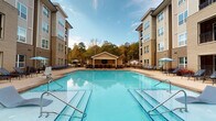Located at 9015 Element Cir in Garner, NC, this outdoor amenity space is centered around a generously sized swimming pool that serves as the focal point of the community's exterior. The pool itself is notably large, with clear blue water, defined lane markings along the bottom, and stainless steel entry railings on multiple sides for easy access. Submerged lounge ledges allow residents to relax partially in the water, adding a resort-style element to the design.

The surrounding deck is finished in a smooth, neutral-toned concrete that provides ample space for lounging and socializing. Numerous chaise lounge chairs and iron-framed seating are arranged throughout the deck, accommodating a variety of outdoor activities. Umbrella-shaded dining tables offer spots for casual gatherings, while raised planters filled with ornamental grasses add a decorative, natural touch to the hardscape.

A charming covered cabana structure anchors the far end of the pool area, providing a shaded communal space with additional seating. The entire courtyard is framed by multi-story brick and siding buildings with private balconies, while a backdrop of mature deciduous trees enhances the sense of privacy and natural ambiance. The overall setting is well-maintained and thoughtfully designed for outdoor recreation and relaxation.