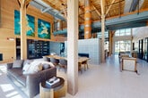 The Great Room at 1 Hanson St in Lynn, MA is a generously sized communal gathering space that masterfully blends architectural warmth with modern design. The interior is defined by soaring ceilings supported by striking exposed timber columns and intricate wooden truss work, creating a dramatic yet inviting atmosphere. Terrazzo flooring runs throughout, reflecting abundant natural light that pours in through large floor-to-ceiling windows and oversized operable glass doors.

The open-plan layout is thoughtfully arranged with distinct seating zones, including lounge areas with plush sofas and accent chairs, communal dining tables, and a bar-height counter with seating. A tiled fireplace serves as a focal point, while curated artwork — including large-scale blue and teal canvases — adds color and character. An upper mezzanine level overlooks the main floor, adding verticality and visual interest.

Large glass doors open directly onto a covered outdoor terrace, which connects to an expansive courtyard featuring a resort-style pool, manicured lawn with Adirondack seating, landscaped garden beds, and a fire pit lounge area. The seamless indoor-outdoor flow and refined finishes make this Great Room an exceptional communal amenity space.