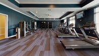 This single-level fitness facility in Fredericksburg, Virginia, is designed to support a comprehensive range of workout activities across several distinct zones. The space is unified by a consistent aesthetic of deep teal-blue walls, crisp white crown molding, and tray ceilings accented with recessed lighting, creating a polished and motivating environment throughout.

The main cardio area is generously proportioned, housing a row of treadmills and elliptical machines along windowed walls fitted with slatted blinds. Wall-mounted televisions are positioned throughout for entertainment during workouts. Natural light filters in through multiple windows, complementing the recessed overhead lighting.

Adjacent to the cardio zone, a dedicated strength training area features a selection of cable and resistance machines arranged along the teal-accented walls, with striped, multi-tone carpet tiles underfoot providing both durability and a refined look.

A separate studio space is enclosed by glass-paned French doors, offering a more intimate setting for group classes or individual training. This room includes floor-to-ceiling mirrors along one wall, a ballet barre, spin bikes, and accessory storage. An additional transitional area with warm wood-look flooring and wainscoting provides a clean, flexible space within the facility.