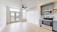 Located at 225 N Sycamore St in Wichita, Kansas, this thoughtfully designed studio offers an efficient and modern single-floor layout that seamlessly combines living and kitchen functions. Light wood-look flooring flows throughout, complementing the crisp white walls and creating a bright, airy atmosphere. The open living area is anchored by a ceiling fan and flooded with natural light from large, tall windows and a set of French doors that open onto a private balcony, offering views of the surrounding Wichita cityscape. A built-in Murphy bed system with integrated white cabinetry and built-in drawers provides smart, space-saving storage while keeping the living area open and versatile. The kitchen is outfitted with slate-blue cabinetry, white subway tile backsplash, and a full suite of stainless steel appliances, including a refrigerator, electric range, over-the-range microwave, and dishwasher. A long hallway leads from the entry to the main living space, with interior doors featuring sleek barn-style hardware. The overall design aesthetic is clean and contemporary, making excellent use of every square foot through intentional built-ins and an open, uncluttered floor plan.