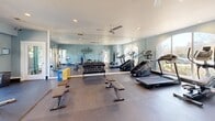 Located in Columbus, Georgia, this fitness center offers a thoughtfully designed exercise space built to support a comprehensive workout experience. The room features light blue walls accented by a bold motivational "WORKOUT" typographic wall mural, creating an energetic and inspiring atmosphere throughout. Large arched and rectangular windows line the walls, flooding the space with natural light and offering views of the surrounding outdoor area, including what appears to be a pool courtyard. A full-length wall mirror expands the sense of space visually while aiding exercisers in monitoring their form. Ceiling fans provide comfortable airflow during intense sessions. The flooring transitions between rubber matting and hard surface zones, catering to a variety of workout types. Equipment is well-distributed and includes treadmills, a stair climber, a rowing machine, and a dual cable functional trainer for strength training. A tiered dumbbell rack stocked with a full range of free weights sits alongside flat workout benches. Medicine balls and stability balls are neatly stored on dedicated racks, and a water cooler ensures easy hydration. Recessed lighting supplements the abundant natural light for consistent illumination at any hour. The space is efficiently organized to maximize usability without feeling crowded.