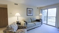 This single-floor residence offers a welcoming and well-appointed living space designed with comfort and functionality in mind. The living room serves as the heart of the home, featuring plush carpeted flooring and a neutral, soft color palette that creates a calm and inviting atmosphere. A generously sized sofa anchors the seating area, complemented by a patterned accent chair that adds a touch of visual interest. Elegant table lamps on either side of the sofa provide warm ambient lighting, enhancing the room's cozy character. A framed botanical artwork adorns the wall, contributing to the understated, tasteful decor. One of the standout features of the living area is the large sliding glass door that floods the space with abundant natural light and opens directly onto a private balcony, offering pleasant views of surrounding greenery. The balcony extends the living space outdoors, providing an ideal spot for relaxation. The room also includes HVAC vents integrated into the ceiling, ensuring year-round climate comfort. Overall, this residence strikes a balance between practical design and a serene, residential ambiance suited to everyday living.