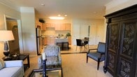 This single-floor residence offers a thoughtfully designed open-concept layout that brings together the main living areas in a cohesive and functional arrangement. The kitchen is fully equipped with stainless steel appliances, including a full-size refrigerator and range, and features warm honey-toned wood cabinetry that extends to the upper walls, providing ample storage. Recessed lighting and under-cabinet fixtures ensure the kitchen is well-lit and practical for everyday use. A raised breakfast bar with bar seating serves as a natural transition between the kitchen and the rest of the living space. Adjacent to the kitchen, a dedicated dining area accommodates a small table and chairs, making it ideal for casual meals. The living area is generously proportioned, with light neutral carpeting that creates a warm and inviting atmosphere. Patterned accent chairs and classic furniture pieces add character to the space. Crown molding details along the upper walls contribute a refined architectural touch throughout. Multiple interior doors suggest additional rooms beyond the main open area, offering privacy and flexibility. The overall ambiance is bright, clean, and welcoming, suited for comfortable day-to-day living.