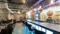 This single-floor bar and lounge space embraces a bold industrial aesthetic, combining raw architectural elements with carefully curated vintage details to create a distinctive atmosphere. The ceiling is left fully exposed, revealing an intricate network of pipes, conduits, and ductwork that adds to the space's gritty, urban character. Pendant lantern-style lights hang at varying heights throughout, casting a warm, ambient glow that contrasts with cool blue accent lighting beneath the bar counter.

The central bar is a standout feature, stretching generously along one side of the room. Its illuminated base emits a striking blue light, lending the counter a modern edge against the otherwise weathered surroundings. Sleek black high-back bar stools line the bar, providing ample seating for guests. Behind the bar, open shelving displays glassware against backlit frosted panels, adding depth and visual interest.

The lounge area features comfortable leather seating arrangements, including armchairs and low tables, offering a relaxed alternative to bar seating. A pool table is visible toward the rear, enhancing the venue's entertainment appeal. Vintage signage, including a notable Standard Oil advertisement, decorates the distressed walls, reinforcing the retro-industrial theme throughout the space.