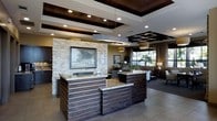 This single-floor space presents a sophisticated and well-appointed lobby and reception area designed to make a strong first impression. At the heart of the space, dual reception desks crafted from rich, dark wood with horizontal slat detailing are topped with elegant granite countertops, offering both style and functionality for guest services. A striking stacked-stone accent wall serves as a dramatic focal point behind the desks, anchoring the space with natural texture and warmth.

The ceiling design is a standout architectural feature, incorporating a combination of dark wood-paneled tray ceilings, geometric coffered sections, and recessed lighting that work together to add depth and dimension. Pendant drum fixtures provide a soft ambient glow that complements the recessed lighting throughout.

Beyond the reception area, a comfortable lounge space extends the welcoming atmosphere, furnished with seating arrangements that invite guests to relax. Large windows allow natural light to filter in, offering views of lush outdoor greenery. The flooring consists of large-format neutral tiles that tie together the warm wood tones and stone textures cohesively. Overall, the space balances a contemporary aesthetic with natural materials, creating a professional yet inviting environment.