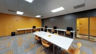 This well-appointed single-floor workspace is designed to support both collaborative and individual work in a dynamic, modern environment. The space is immediately defined by its bold color scheme — striking amber-yellow accent walls contrast against cool charcoal-gray surfaces, creating an energetic yet focused atmosphere throughout.

The layout is thoughtfully organized into distinct functional zones. A central white rectangular meeting table surrounded by ergonomic amber-toned chairs serves as the primary collaboration hub, ideal for group discussions or team sessions. Along the perimeter, continuous white L-shaped countertop workstations provide ample desk space for individual computing, with multiple desktop computers and chairs positioned at regular intervals to accommodate several users simultaneously.

A shared office equipment station, including a multifunction printer, is conveniently accessible within the space, supporting day-to-day operational needs. A display screen mounted near the workstations adds a presentational element to the room. Entry is provided through a distinctive frosted glass door with warm orange-toned framing, adding a refined architectural detail.

The flooring features a patterned commercial carpet with interlocking gray and gold geometric tiles, tying together the room's color palette cohesively. Overhead, a suspended tile ceiling with recessed panel lighting ensures consistent, even illumination throughout the entire space.