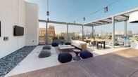 Located in the heart of Los Angeles' vibrant Arts District, this contemporary rooftop space at 695 S Santa Fe Ave offers a striking blend of modern design and urban outdoor living. The space is defined by clean white walls, polished concrete flooring, and thoughtfully integrated landscaping, including dramatic desert cacti and lush planters that add organic texture to the otherwise minimalist aesthetic.

A spacious open-air lounge area serves as the centerpiece, furnished with a plush white sofa, casual floor poufs, and a low coffee table arranged atop a patterned area rug. String lights overhead create an inviting atmosphere, while a patio heater ensures year-round comfort. An adjacent dining area features a wooden table and modern chairs, ideal for gathering and entertaining.

A partially enclosed corner area with soaring walls houses a wall-mounted flat-screen TV, decorative river rock ground accents, and a raised wood platform, creating a semi-private retreat within the open rooftop. Floor-to-ceiling glass panels and steel railings frame sweeping views of the Downtown Los Angeles skyline, flooding the space with abundant natural light. A pergola-style steel overhead structure ties the entire space together, reinforcing the industrial-modern character that defines this exceptional urban rooftop.