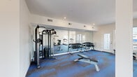 Located at 71 Connecticut Ave in Norwalk, Connecticut, this fitness center offers a well-appointed, single-floor exercise space designed to support a comprehensive workout experience. The open layout provides ample room to move freely, with patterned blue and gray carpet tiles covering the floor throughout, lending a clean and modern aesthetic.

The space is equipped with multiple treadmills, an elliptical, and a stationary bike, all positioned near large windows that flood the area with natural light and offer views of the surrounding neighborhood. A multi-station cable strength machine anchors the strength training zone, complemented by a full dumbbell rack with a wide range of weights and an adjustable workout bench.

Floor-to-ceiling mirrors line one wall, enhancing the sense of openness and allowing users to monitor their form during exercise. Structural columns are integrated throughout the space and are finished in crisp white, maintaining a cohesive, contemporary look. Recessed lighting provides consistent illumination throughout, while HVAC vents ensure proper climate control. Multiple entry points, including exterior-facing doors, contribute to easy access and good ventilation. The clean white walls and bright natural light create an energizing and welcoming environment well-suited for a wide range of fitness activities.