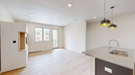 This well-designed one-bedroom, one-bathroom unit offers a thoughtfully efficient layout across a single floor. The open-concept kitchen and living area form the heart of the space, where light wood-style flooring, white walls, and recessed lighting create a bright, clean aesthetic throughout.

The kitchen is a standout feature, anchored by a large central island with granite countertops, an undermount sink, and dark cabinetry below. Stainless steel appliances—including a refrigerator, electric range, and microwave—complement the white upper cabinetry and tile backsplash. Stylish dome pendant lights with gold interiors add a modern accent overhead.

The living area is open and airy, benefiting from natural light through large windows and a glass door that leads to a private balcony overlooking the surrounding neighborhood. A built-in media wall with a TV mount and wood-accented shelving provides a functional focal point.

The spacious primary bedroom features a ceiling fan, a bright window, and light wood flooring. The bathroom is well-appointed with a soaking tub, marble-style tiled surround, granite vanity countertop, and wood cabinetry. A dedicated walk-in closet is directly accessible from the bathroom.