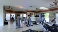 This fitness center is a dedicated, single-floor workout facility designed to support a well-rounded exercise routine. The space is outfitted with a variety of cardio machines, including multiple treadmills and elliptical trainers, arranged to allow comfortable movement between equipment. A weight bench is also present, catering to strength training needs. The interior features a neutral color palette accented by a bold red border near the crown molding, adding a dynamic visual element to the room. Tile flooring throughout provides a durable and easy-to-maintain surface suitable for high-traffic use. Overhead, multiple ceiling fans ensure consistent air circulation, while track lighting supplements the ample natural light streaming in through large windows and a skylight. The windows offer views of surrounding greenery, contributing to an open and refreshing atmosphere. Decorative artwork on the wall adds a touch of character to the functional space. French doors at one end of the room enhance accessibility and further connect the interior to the outdoors. Overall, the fitness center is bright, spacious, and thoughtfully laid out to accommodate a range of workout activities.